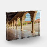 Stanford University Photo Block<br><div class="desc">The image showcases a long row of stone arches and columns, creating a corridor-like structure. The arches frame a courtyard with buildings featuring red-tiled roofs and lush greenery. The floor is tiled in a checkered pattern of beige and light brown. The architecture is classical, with detailed carvings on the column...</div>