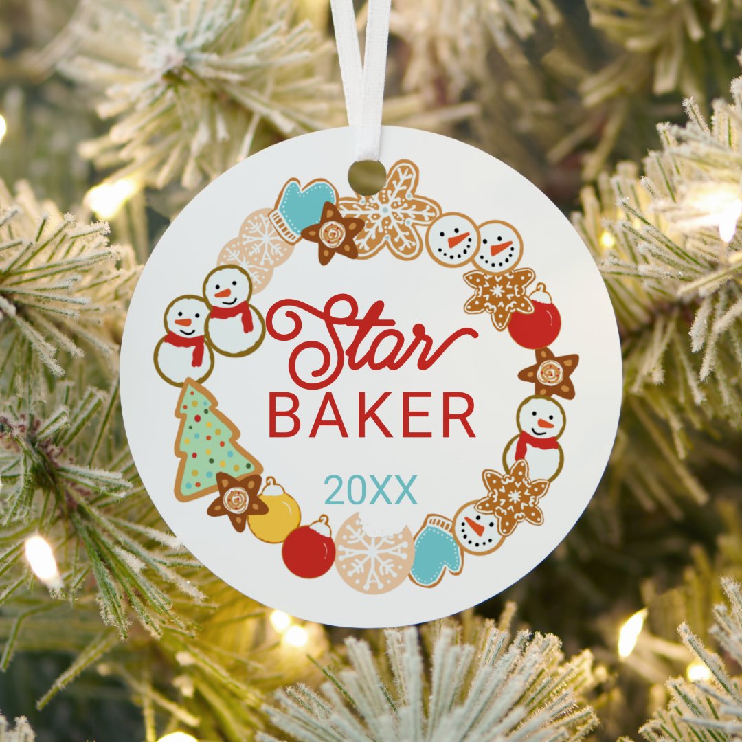 A round white metal ornament with a wreath of illustrated Christmas cookies, including snowmen, stars, mittens, and a decorated tree, hanging from a white ribbon on a frosted, lit Christmas tree.