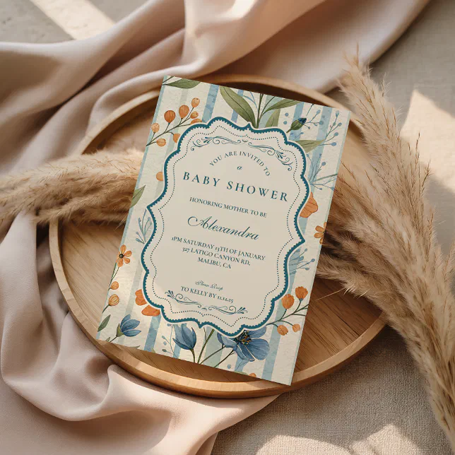 A mixed pattern baby shower invitation is displayed on top of pampas reeds, which are placed on a wooden bowl and a pink-beige satin fabric.