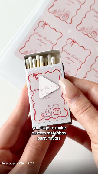 A hand holds an open matchbook-style favor box with pink scalloped design. Close-up, tactile, boutique-craft mood. 