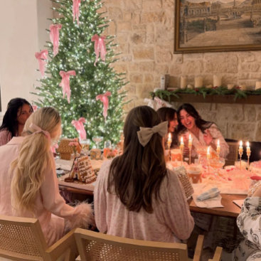 A group gathered at a long candlelit dinner table, seen from behind, with a Christmas tree tied in soft pink bows glowing in a stone-walled room. Warm, intimate, feminine holiday mood.