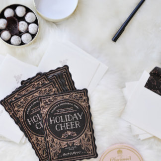 Dark ornate cards labeled with foil-style script lie among scattered envelopes and ribbon spools on a light surface — moody, glam, tactile.
