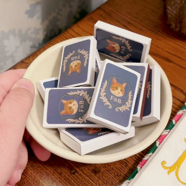 Small illustrated matchboxes scattered on a pale dish atop a wooden table with gold-accented gift wrap. Delicate, artisanal, small-batch favor styling.