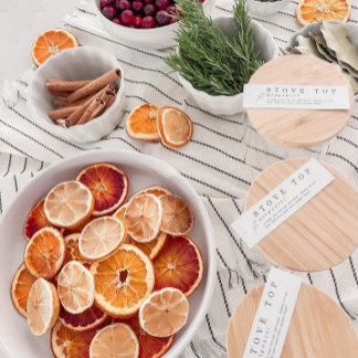 Dried citrus slices, cinnamon sticks, cranberries, and pine sprigs surround wooden jar-lid labels reading "Enjoy Toff..." on a striped linen cloth. The tone is rustic and handmade—ideal for DIY holiday gifting.