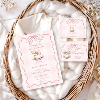 A baby shower invitation and matching baby clothing arranged in a woven basket with soft fabric. The styling feels cozy and coordinated with a soft, delicate theme.