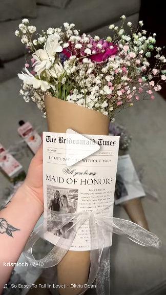 A hand holds a tall kraft bouquet wrapped in a newsprint-style proposal flyer, topped with baby's breath and deep-red blooms. Romantic, thoughtful, gift-reveal moment.