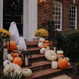 Ghost figures and a pumpkin-lined staircase decorate a brick porch entrance at dusk. The tone is spooky and charming.