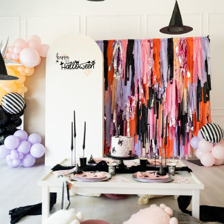 A whimsical dessert table with fringed streamer backdrop in orange, pink, and black, styled with balloons and a witch hat — playful, spooky-chic, festive.
