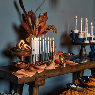A lit menorah stands on a wooden console styled with dried leaves and pumpkins against a dark wall. The tone is warm and ceremonial.