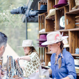 An outdoor scene with guests engaging in custom art or sketching experiences. Warm lighting, hats, and rustic props evoke a handcrafted, personal mood. The environment feels lively and casual. It highlights artisanal, guest-centered interactivity.