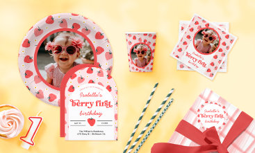 The image showcases a delightful "Berry First" birthday celebration with a charming strawberry theme. A pink and red color palette runs throughout, with playful strawberry patterns adorning the plates, cups, napkins, and invitations. A custom photo plate and napkins feature the birthday child’s smiling face, bringing a personal touch to the setup. Sweet details like soft pink cupcakes, a number one candle, striped paper straws, and a gingham-wrapped gift complete the festive scene, all set against a warm yellow background that enhances the cheerful atmosphere.