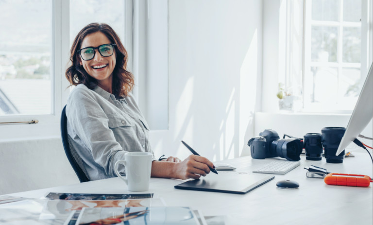 A smiling woman in glasses and a chambray shirt sits at a bright, sunlit white desk with a coffee mug, camera gear, and a drawing tablet. Airy, natural-light workspace with a relaxed, professional feel. 