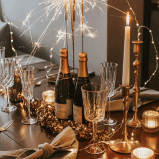 Champagne bottles, sparklers, and gold confetti surround glassware on a dark table setting. The tone is glamorous and celebratory.