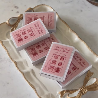 A set of blush pink personalized matchboxes displayed in a cozy kraft basket. The styling is minimal but meaningful, with a vintage touch.