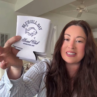 A person holds up a branded coffee mug with a nautical logo toward the camera in a bright indoor setting — friendly, personal, casual.
