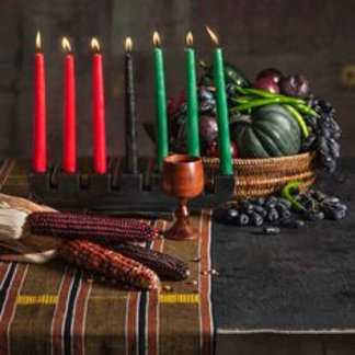 Red and green candles stand among corn husks, gourds, and woven fabric on a dark table. The tone is earthy and symbolic.