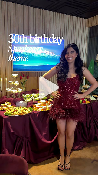A person in a burgundy fringe dress poses at a speakeasy-themed birthday party with a dessert table and beach backdrop screen. Festive, glamorous, event-lifestyle shot.