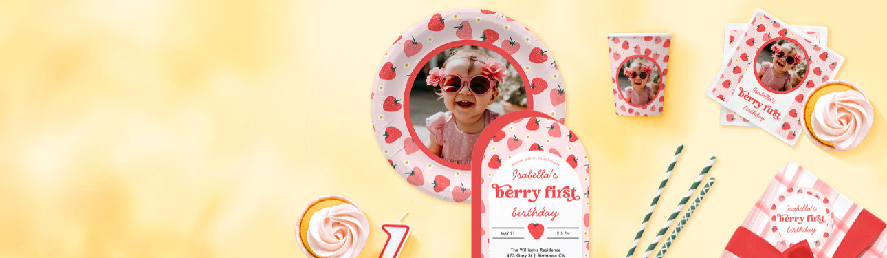 An array of colorful strawberry themed first birthday part supplies and invitations