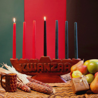A bold red background with a lit kinara (candle holder), fruit, and Kwanzaa symbols arranged in a cultural display. The look is rich, meaningful, and affirming.