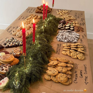 A rustic spread of cookies and lit red taper candles rests atop a kraft-paper backdrop nestled in greenery — homey, warm, abundant.