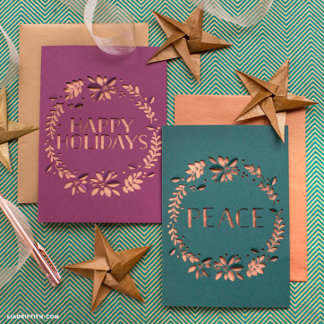 Plum and slate-blue cards with gilded foliage lettering lean together atop a green herringbone surface, flanked by wooden star ornaments and copper ribbon — rich, festive, artisanal warmth.