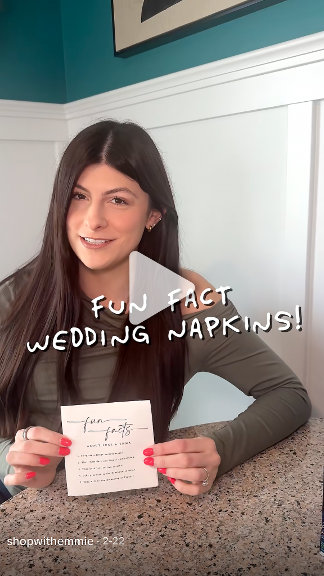 A dark-haired person holds up custom napkins to the camera in a home setting. Casual, personable, creator-selfie style. 
