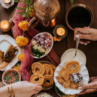 An overhead spread of fried snacks, curries, and sweets sits on a dark table as a hand reaches in. The tone is rich and communal.