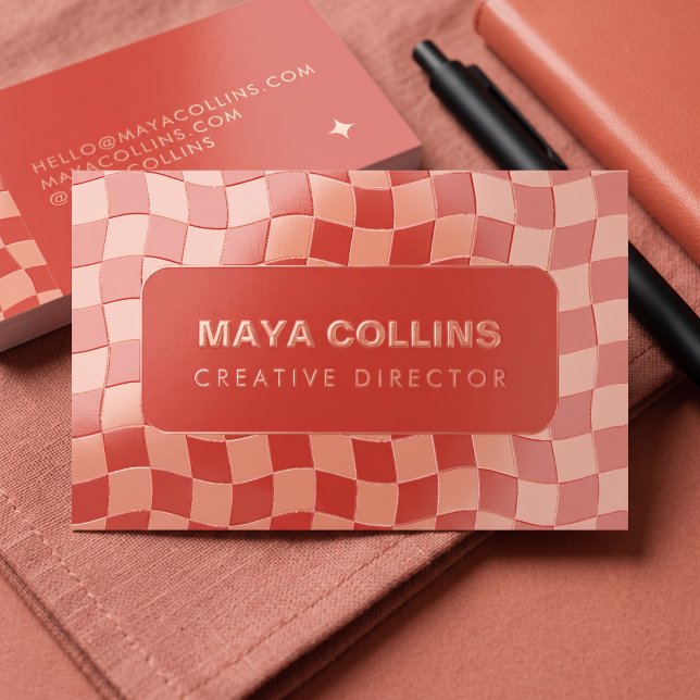 A trendy red and pink wavy checkerboard business card featuring rose gold foil typography sitting alongside a black pen resting on a terracotta-colored fabric.