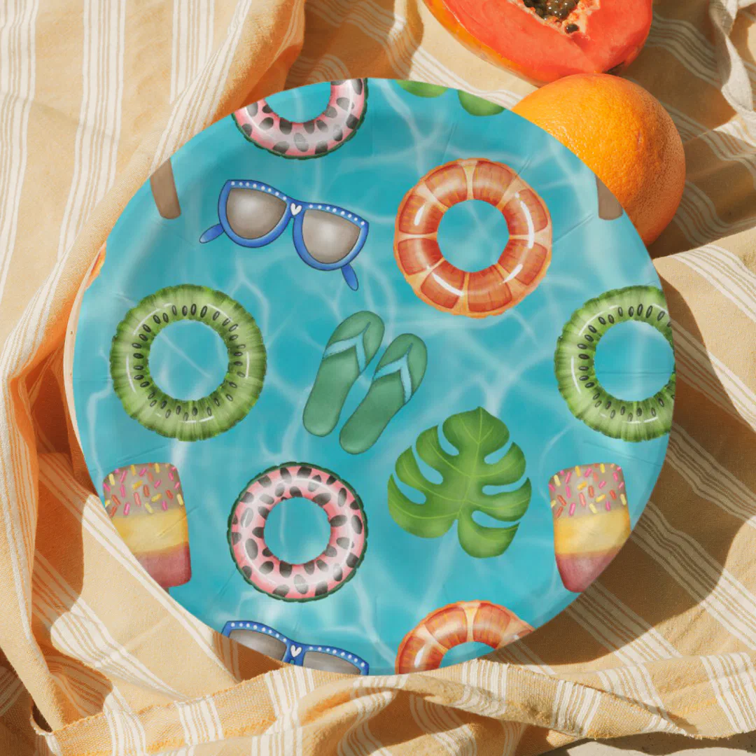 A circular party plate featuring a bright blue rippling pool water print scattered with playful summer motifs including kiwi, watermelon, and orange-slice ring floats, a pair of blue-framed sunglasses, green flip flops, a tropical monstera leaf, and colorful popsicles, photographed on a yellow striped beach towel with fresh tropical fruit in the corner.
