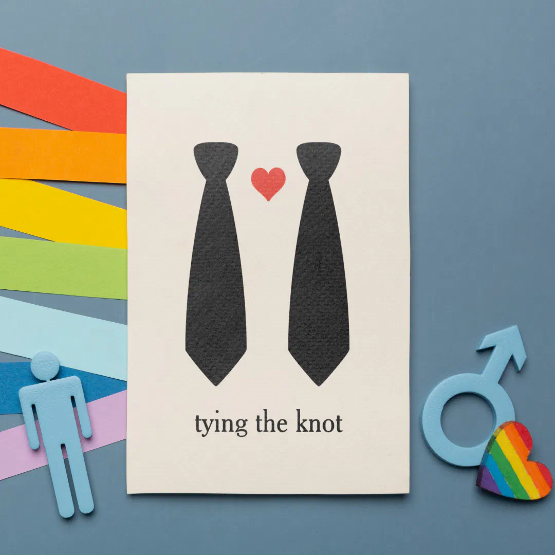 A cream wedding invitation card propped against a blue-gray surface, surrounded by layered rainbow paper strips, a small blue human figure, a male gender symbol, and a rainbow heart. The card features two illustrated black neckties with a small red heart between them.