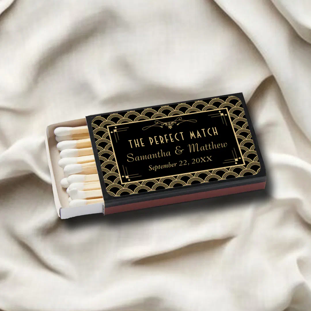Art deco wedding favor matchboxes in black and gold with a scalloped pattern, one box open to show white-tipped matches, resting on cream satin fabric.