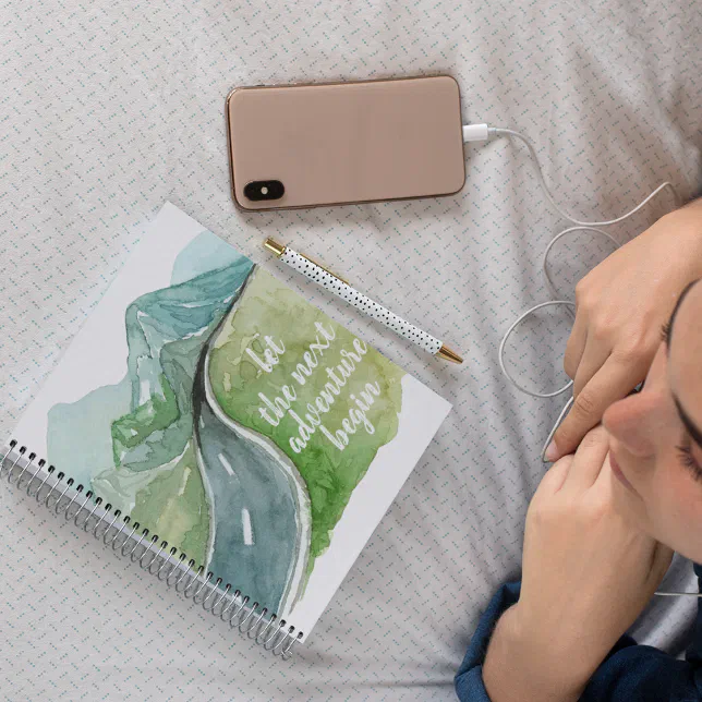 A notebook rests on a bedspread, with  a pen and a phone beside it. A person is seen at the corner of the picture looking at the notebook.