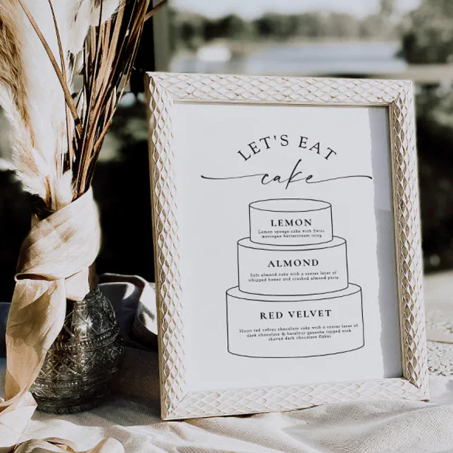 A white cake sign with a cake graphic in a white frame sitting next to a vase with some floral accents.