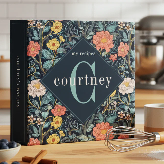 A personalized recipe binder set on a kitchen counter with a whisk, a mug, a blueberry-filled bowl, and some cinnamon sticks seen in the front, and a blurred mixer seen in the background.