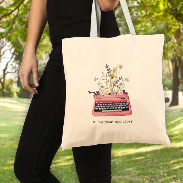 Write your own story, Writers Tote Bag (A woman holding a tote bag with a typewriter graphic and wildflowers & text: "write your own story")
